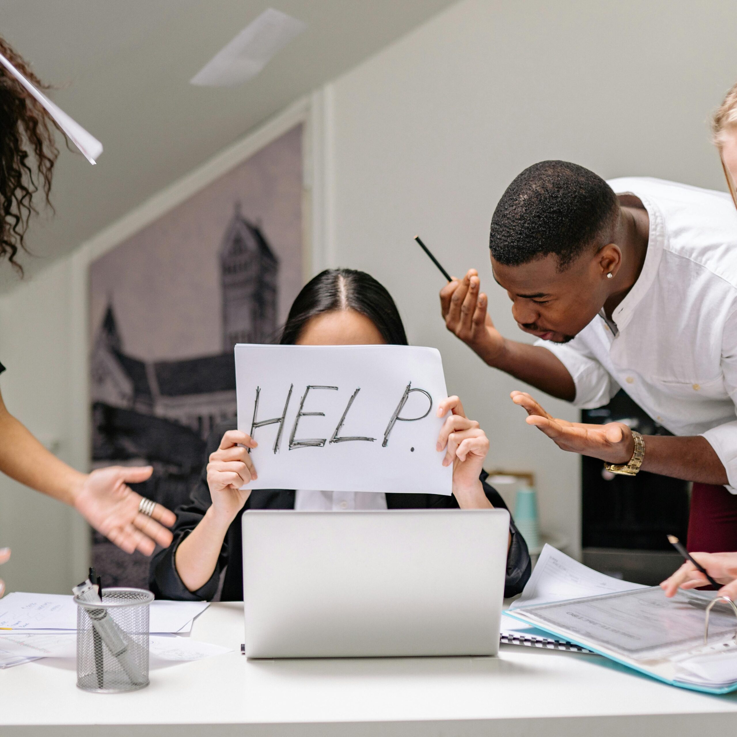 A stressed employee holds a 'HELP' sign at a busy office desk, depicting workplace pressure.