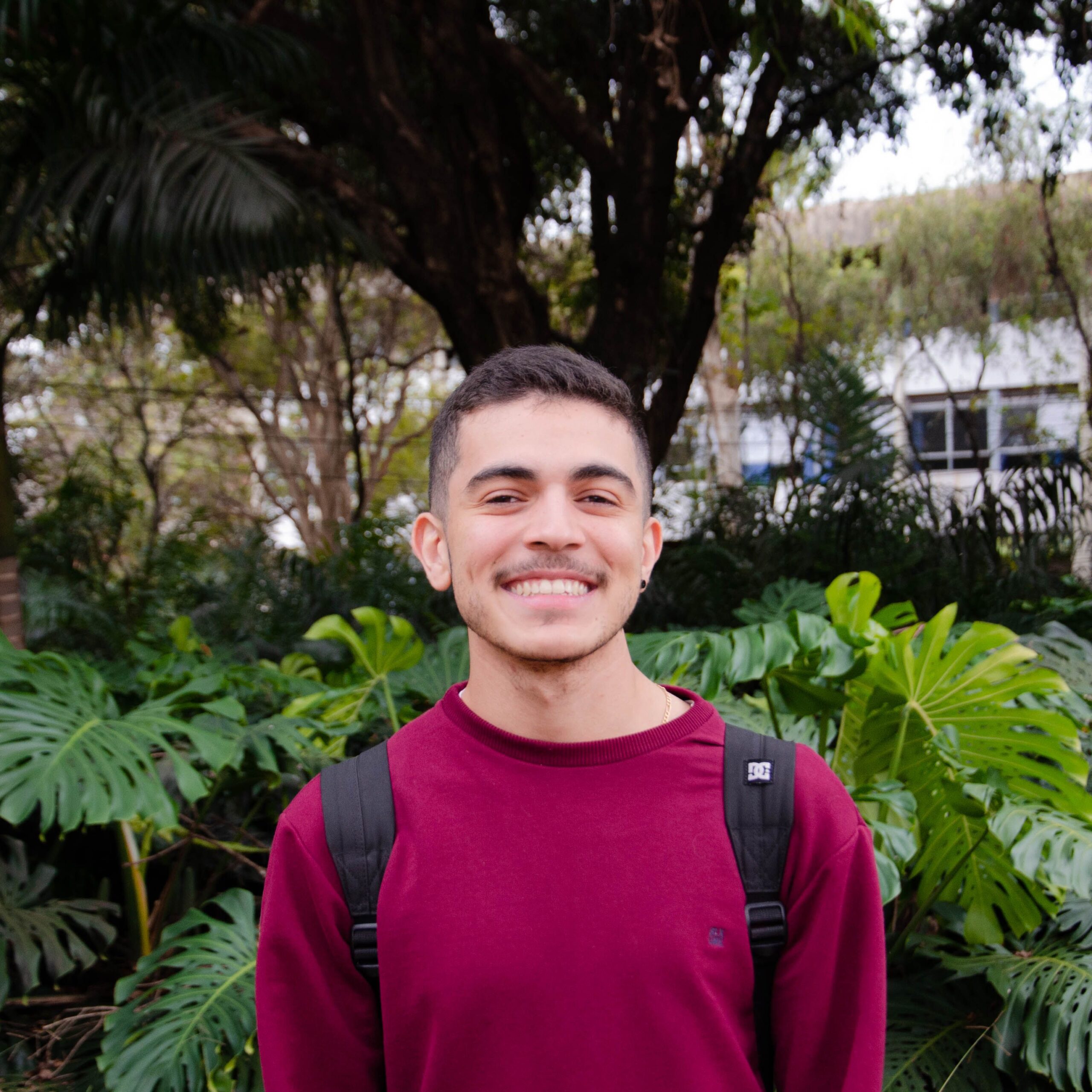 Cheerful young man wearing a backpack, standing in front of lush foliage outdoors.